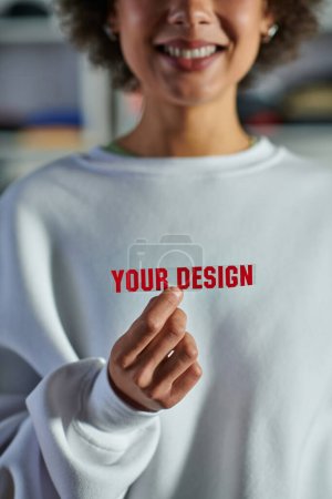 Cropped view of smiling blurred african american designer in sweatshirt holding printing layer with your design lettering in print studio, focused business owner managing workshop