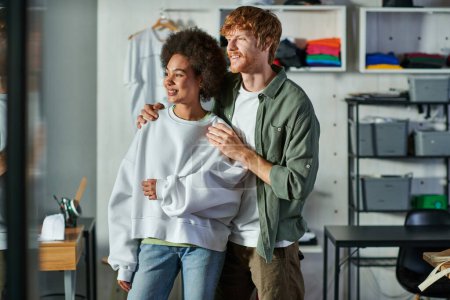 Cheerful young redhead artisan hugging african american colleague and looking away while standing in blurred print studio, collaborative business owners working together 