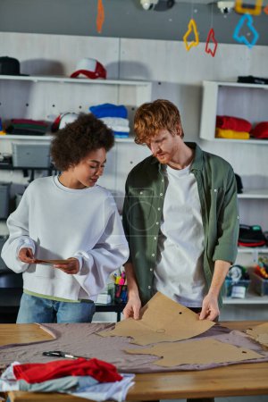 Photo for Young redhead craftsman holding sewing pattern near cloth while working with african american colleague in blurred print studio, collaborative business owners working together - Royalty Free Image
