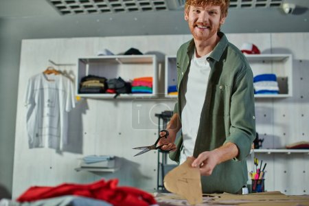 Smiling redhead craftsman looking at camera while holding scissors and sewing pattern near fabric on table in print studio, multitasking business owner managing multiple project
