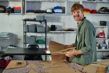 Smiling young redhead craftsman looking at camera while holding sewing patterns near scissors and fabric on table in print studio, multitasking business owner managing multiple project