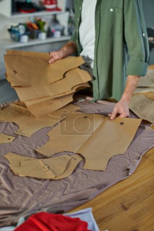 Vista recortada de artesanos sosteniendo patrones de costura cerca de la tela en la mesa mientras trabajan en un estudio de impresión borrosa, dueño de un negocio multitarea administrando múltiples proyectos