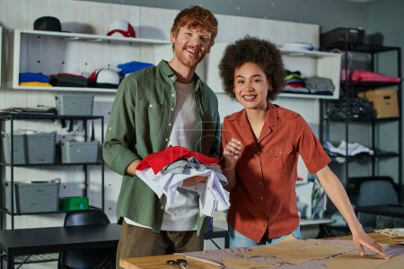 Photo for Positive young multiethnic craftspeople looking at camera while working with clothes, fabric and sewing patterns in print studio at background, ambitious young entrepreneurs concept - Royalty Free Image