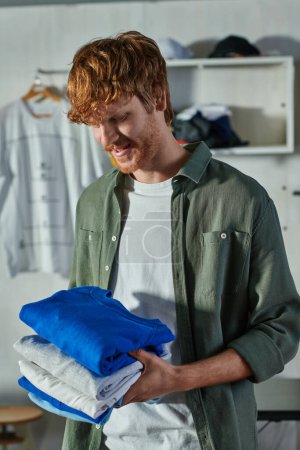Cheerful young redhead craftsman holding clothes while standing and working in blurred print studio at background, customer-focused entrepreneur concept 