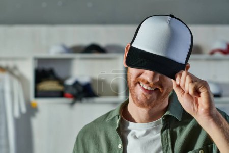 Photo for Cheerful young craftsman smiling and covering face with snapback while standing and working in blurred print studio at background, customer-focused entrepreneur concept - Royalty Free Image