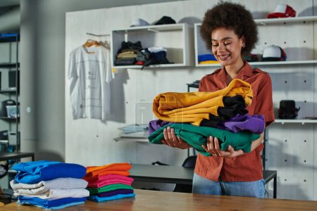 Positive junge afrikanisch-amerikanische Handwerkerin mit Kleidung am Arbeitstisch, während sie im verschwommenen Druckstudio im Hintergrund steht, kundenorientiertes Unternehmerkonzept 