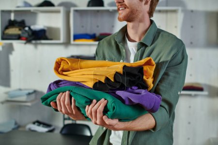 Cropped view of smiling young redhead craftsman holding clothes while working and standing in blurred print studio at background, customer-focused entrepreneur concept 