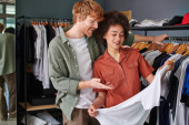Smiling young redhead craftsman pointing at clothes on hanger and talking to african american colleague in blurred print studio, young small business owners concept  Poster #664661744