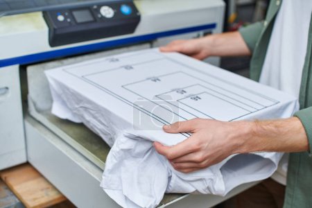 Cropped view of young craftsman holding t-shirt with marking near screen printing machine and working in print studio, customer-focused small business concept