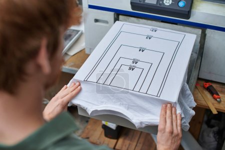 Hohe Blickwinkel auf junge verschwommene Handwerker, die mit T-Shirt mit Markierung und Siebdruckmaschine im Hintergrund im Druckstudio arbeiten, kundenorientiertes Kleinunternehmenskonzept