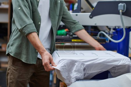 Cropped view of young craftsman holding t-shirt with marking while working with screen printing machine in print studio at background, customer-focused small business concept