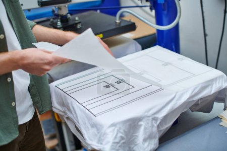 Cropped view of young artisan holding printing layer near t-shirt with marking and working with screen printing machine in print studio, customer-focused small business concept