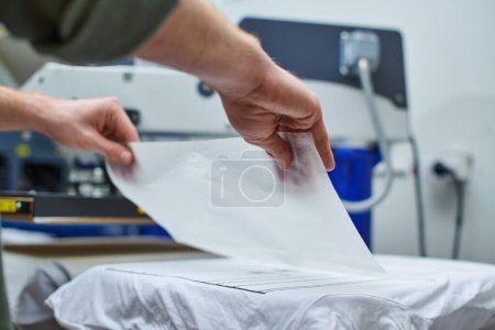 Téléchargez les photos : Vue recadrée du jeune artisan mettant la couche sur le t-shirt et travaillant sur la machine de sérigraphie en arrière-plan dans le studio d'impression, concept de petite entreprise axé sur le client - en image libre de droit