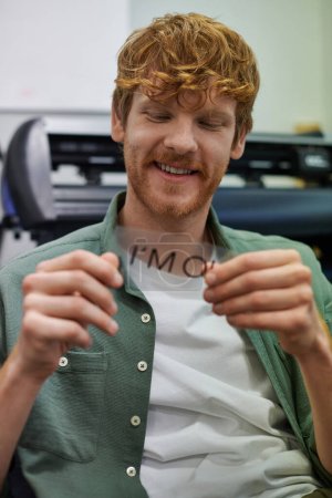 Photo for Smiling young redhead craftsman holding layer with I'm ok lettering while working near screen printing machine in workshop, small business owner working on project - Royalty Free Image