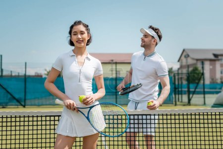 szczęśliwa kobieta w aktywnym ubraniu trzyma rakietę tenisową w pobliżu mężczyzny, tenisiści na korcie