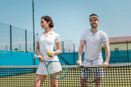 szczęśliwy mężczyzna i kobieta w odzieży sportowej stojący z rakietami tenisowymi na korcie, fitness i zdrowie