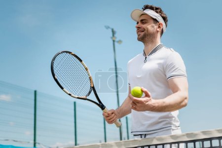 cheerful tennis player in visor cap holding racket and ball on court, fitness and motivation, joy
