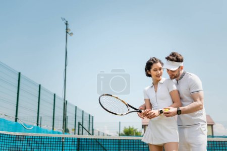 happy man teaching girlfriend how to play tennis on court, holding rackets and ball, sport and fun
