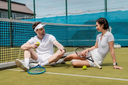 Foto de Feliz hombre y mujer en ropa deportiva sentado en la cancha de tenis, raquetas y pelotas, verano, deporte de pareja - Imagen libre de derechos