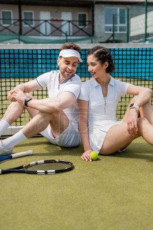 Téléchargez les photos : Couple positif assis près du filet de tennis, raquettes et ballon, activité estivale, loisirs et plaisir - en image libre de droit
