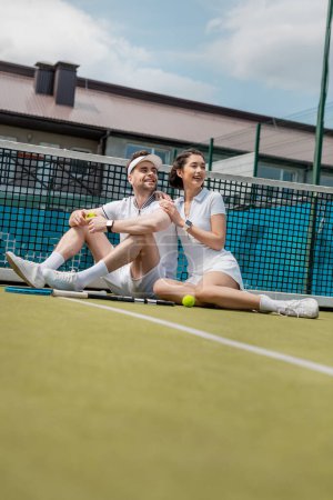 cheerful man and woman sitting near tennis net, rackets and ball, summer sport, leisure and fun