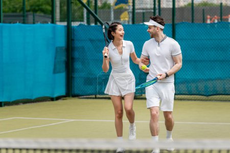 happy sporty couple in active wear walking on tennis court, rackets and ball, hobby and sport