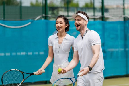 fröhliches Paar in aktiver Kleidung, lachend auf Tennisplatz, Spieler, Schläger und Ball, Sport