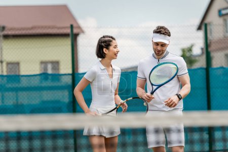 wesoły mężczyzna w aktywnym ubraniu patrząc na rakietę tenisową, szczęśliwa kobieta uśmiechnięta na korcie tenisowym, sport
