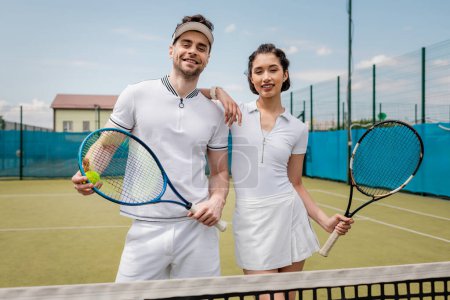 szczęśliwa sportowa para w aktywnym ubraniu patrząc na kamerę na korcie tenisowym, hobby i sport, lato