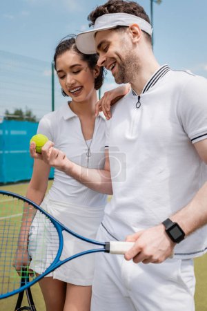 cheerful couple in active wear looking at tennis ball on court, leisure and sport, summer fun
