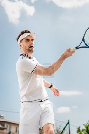przystojny mężczyzna w visor czapka i aktywne nosić rakietka trzyma na kort tenisowy, backhand, hobby i sport