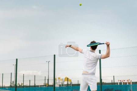 Photo for Backhand, man in active wear playing tennis, holding racket, hitting ball, backhand, back view - Royalty Free Image