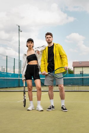 man and woman in stylish active wear standing on court with tennis rackets, healthy lifestyle, sport