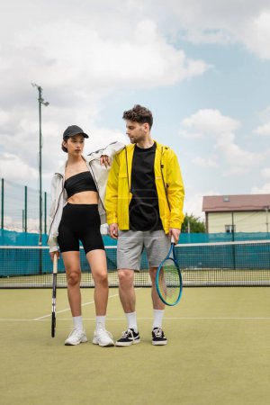Téléchargez les photos : Couple à la mode debout sur un court de tennis avec raquettes, vêtements de sport élégants, joueurs de tennis - en image libre de droit