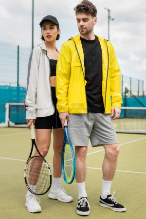 sport, fashionable couple standing on tennis court with rackets, man and woman in stylish outfits