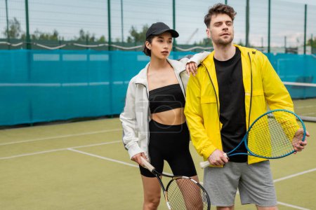deporte, pareja de moda de pie en la cancha con raquetas de tenis, hombre y mujer en trajes elegantes