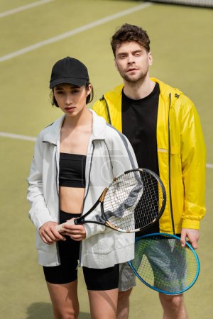 sport i styl, modny mężczyzna i kobieta w odzieży sportowej trzyma rakiety tenisowe na korcie
