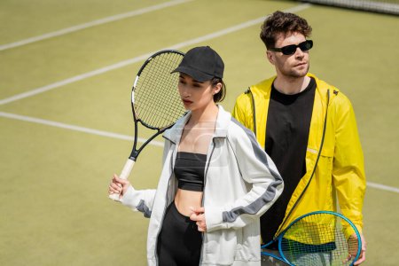 Foto de Pareja deportiva de moda, hombre en gafas de sol y mujer en gorra con raquetas en la pista de tenis - Imagen libre de derechos