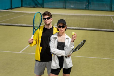 moda i sport, mężczyzna w okularach przeciwsłonecznych i kobieta w czapce trzyma rakiety i piłkę na korcie tenisowym