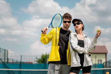 moda i sport, stylowa para w okularach przeciwsłonecznych, trzymając rakiety i piłkę na korcie tenisowym