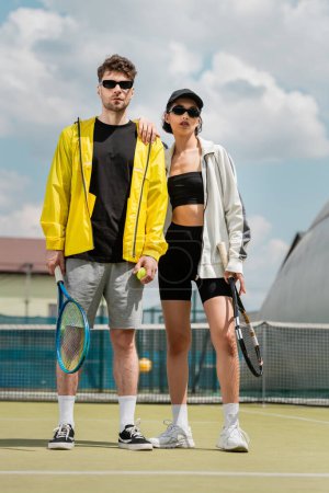 hobby i sport, stylowy mężczyzna i kobieta w okularach przeciwsłonecznych trzymających rakiety i piłkę na korcie tenisowym