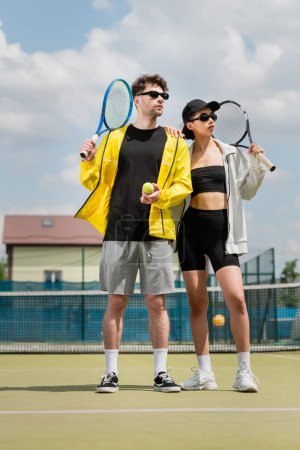 motywacja i moda sportowa, mężczyzna i kobieta w okularach przeciwsłonecznych trzymających rakiety i piłkę na korcie tenisowym