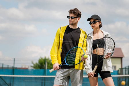 sport letni, mężczyzna i kobieta w okularach przeciwsłonecznych stojący na korcie tenisowym z rakietami, moda sportowców