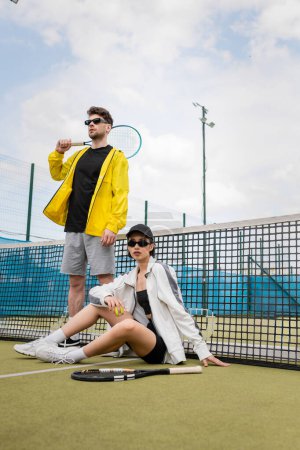 hobby and sport, man and woman in sunglasses posing near tennis net with rackets, sporty fashion