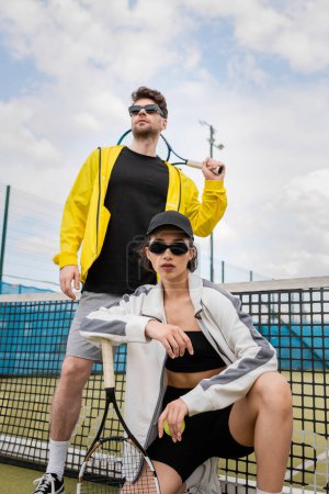 Mann und Frau mit Sonnenbrille posieren in der Nähe des Tennisnetzes mit Schlägern, sportliche Mode, Sport als Hobby,