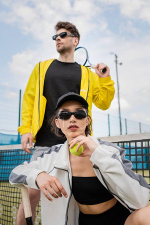 woman in sunglasses and cap posing near man with tennis racket, sporty fashion, sport as a hobby