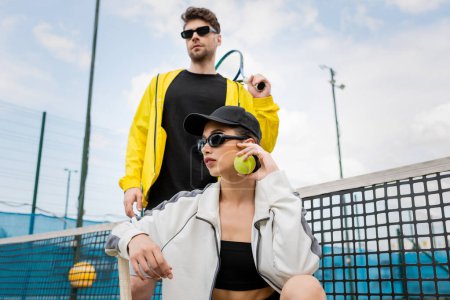 kobieta w okularach przeciwsłonecznych i czapce pozowanie w pobliżu sportowca z rakietą tenisową, moda na aktywne noszenie