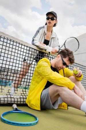 sportowa para w okularach przeciwsłonecznych w pobliżu siatki tenisowej, trzymanie rakietek i piłki, aktywna moda na zużycie