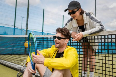 glücklicher Mann und Frau in aktiver Kleidung, die sich in der Nähe des Tennisnetzes auf dem Platz ausruhen, Sportbekleidung Mode, Sport