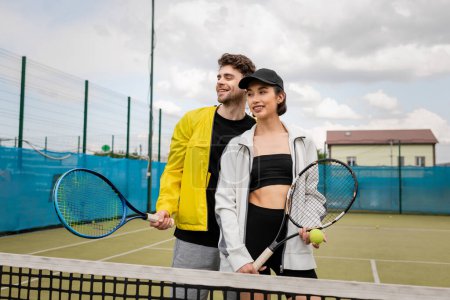 szczęśliwy mężczyzna i kobieta w aktywnym ubraniu trzymając rakiety w pobliżu sieci na korcie tenisowym, styl życia, uśmiech